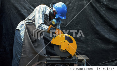 Worker using yellow metal cutting saw while wearing blue hard hat gloves apron ear protection and respirator mask Worker using yellow metal cutting saw while wearing blue hard hat gloves apron ear protection and respirator mask 130301847