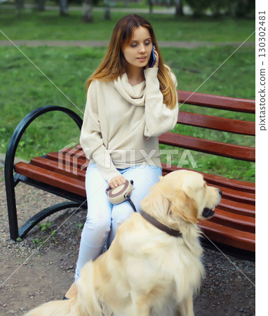 Young woman owner talking on phone with Golden Retriever dog sitting on bench in summer park 130302481