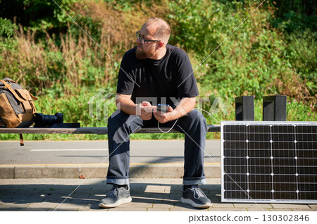 Bearded man using smartphone connected to photovoltaic solar panel. Integration of sustainable renewable energy into everyday life, demonstrating practical use of solar power for charging devices. 130302846