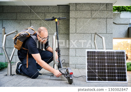 Man connects solar panel to electric scooter for charge, highlighting practical application of sustainable renewable energy. Concept of integration of eco-friendly technology in urban transportation. 130302847
