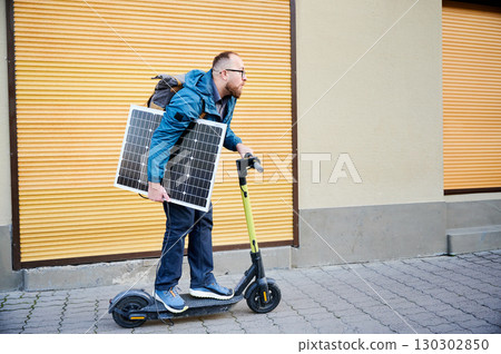 Man rides electric scooter while carrying solar panel. Integration of solar power as sustainable energy source for charging electric scooters, promoting eco-friendly urban transportation. 130302850
