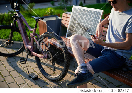 Man cyclist using solar panel for charging electric mountain bike outdoors. Guy in sunglasses sits on bench, holding phone, casually dressed in t-shirt, shorts, enjoying sunny outdoor setting. Man cyclist using solar panel for charging electric mountain bike outdoors. Guy in sunglasses sits on bench, holding phone, casually dressed in t-shirt, shorts, enjoying sunny outdoor setting. 130302867
