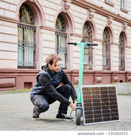 Man connects solar panel to electric scooter for charge, highlighting practical application of sustainable renewable energy. Concept of integration of eco-friendly technology in urban transportation. 130302872