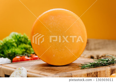 Whole round cheese wheel on wooden board with herbs and vegetables on orange background Whole round cheese wheel on wooden board with herbs and vegetables on orange background 130303011