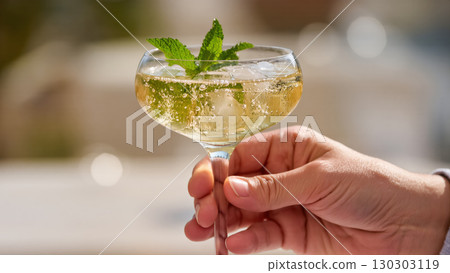 Elegant Hugo cocktail with mint and ice in coupe glass, held by hand in outdoor setting, refreshing sparkling summer drink 130303119