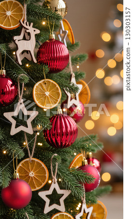 Close-up of Christmas tree decorated with red baubles, wooden ornaments, and dried orange slices in warm festive lighting. 130303157