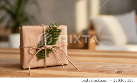 Eco-friendly Christmas gift wrapped in kraft paper with twine and pine branch, placed on wooden table in cozy home interior. 130303195