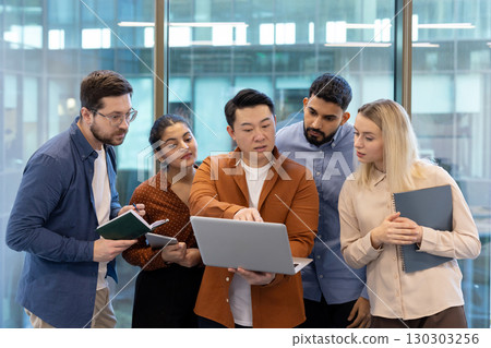 A diverse team collaborates around a laptop in a modern office setting, brainstorming ideas and strategies. 130303256