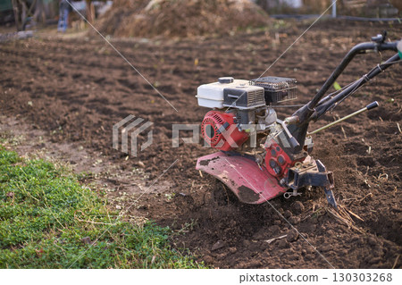 Farming equipment tilling soil in a rural field with red cultivator and green 130303268