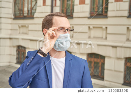 Caucasian male adult adjusting glasses wearing face mask in blue suit outdoors Caucasian male adult adjusting glasses wearing face mask in blue suit outdoors 130303298