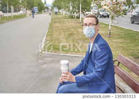 Caucasian male adult sitting on park bench wearing mask holding coffee on street Caucasian male adult sitting on park bench wearing mask holding coffee on street 130303313