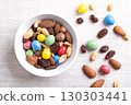 Rainbow trail mix in a white bowl on linen fabric. A snack mix of candy coated milk chocolate peanuts, roasted peanuts, raisins, and roasted almonds. A combination of sweets, dried fruits, and nuts. 130303441