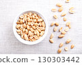 Roasted peanuts in a white bowl on linen fabric. Ready-to-eat crunchy snack food made from fruits of Arachis hypogaea. Light golden snack nuts with natural, nutty aroma, and firm, crunchy texture. 130303442