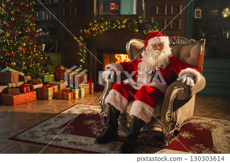 Merry Christmas. Happy Santa Claus sitting in chair in festive living room with fireplace decorations Christmas tree. Santa in cozy home on Christmas eve. Winter New Year Holiday Celebrations 130303614