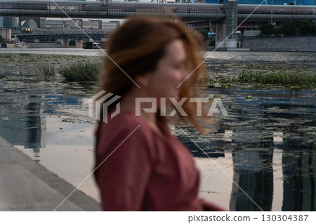Blurred woman in a red dress sits by the Moscow River with the futuristic Bagration Bridge and modern city buildings in the background Blurred woman in a red dress sits by the Moscow River with the futuristic Bagration Bridge and modern city buildings in the background 130304387