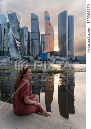 Woman in a red dress sits by the Moscow River with the iconic skyscrapers of Moscow City in the background Woman in a red dress sits by the Moscow River with the iconic skyscrapers of Moscow City in the background 130304389