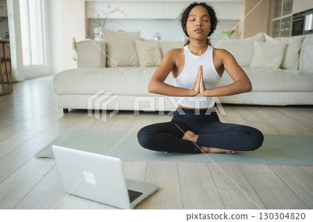 Yoga mindfulness meditation online. African woman practicing yoga with online lessons in laptop at home. Woman sitting in lotus pose meditating relaxing. Girl doing breathing practice. Yoga at home 130304820