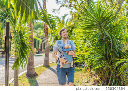 Young father walks in nature with his baby son nestled close in a parent-facing ergonomic baby carrier. Bonding, warmth, and modern parenting concept in a peaceful outdoor setting 130304864