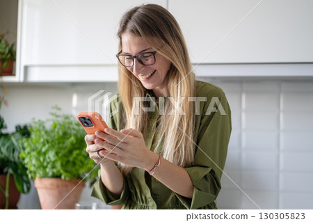 Positive woman typing messages or video calling on smartphone standing in home kitchen with smile, 130305823