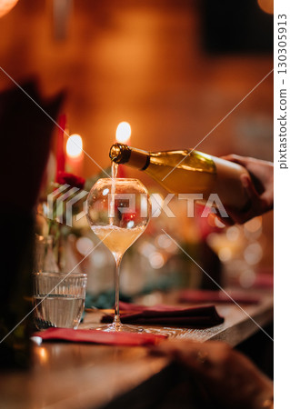 Hand pouring white wine into glass at festive dinner table with candlelight and decor 130305913