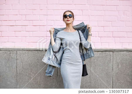 Modern urban woman posing in denim jacket and sunglasses against pastel pink wall. Street fashion portrait with casual elegance and trendy. Perfect for social media, advertising, lifestyle content Modern urban woman posing in denim jacket and sunglasses against pastel pink wall. Street fashion portrait with casual elegance and trendy. Perfect for social media, advertising, lifestyle content 130307317