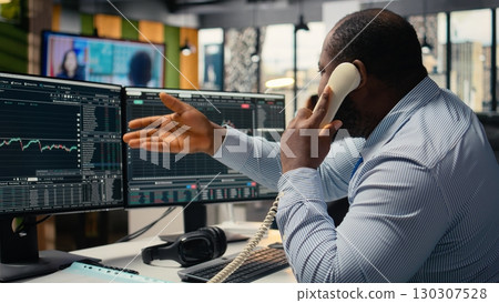 Distressed brokerage firm trader making phone calls to clients in response to Black Swan event shock. African american man evaluate stock exchange impact of unexpected recession Distressed brokerage firm trader making phone calls to clients in response to Black Swan event shock. African american man evaluate stock exchange impact of unexpected recession 130307528