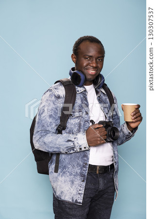 Smiling photographer with headphones around his neck stands against blue background, holding both his DSLR camera and coffee cup. He portrays readiness and professionalism in his photography career. Smiling photographer with headphones around his neck stands against blue background, holding both his DSLR camera and coffee cup. He portrays readiness and professionalism in his photography career. 130307537