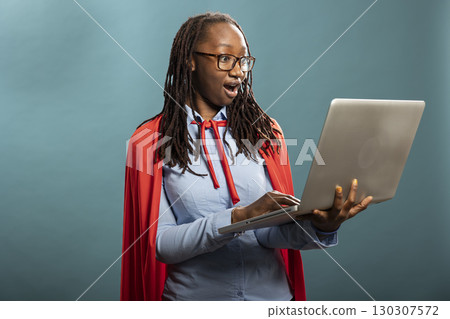 Shocked african american woman in a red cape stares at her laptop with wide eyes. Black female model with expressive reaction conveying excitement and unexpected positive news. Shocked african american woman in a red cape stares at her laptop with wide eyes. Black female model with expressive reaction conveying excitement and unexpected positive news. 130307572