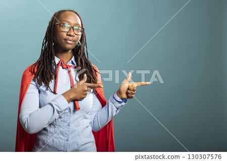 African American woman in red superhero cape gestures to side with index fingers, standing against isolated background with neutral expression. Serious black female individual pointing sideways. African American woman in red superhero cape gestures to side with index fingers, standing against isolated background with neutral expression. Serious black female individual pointing sideways. 130307576