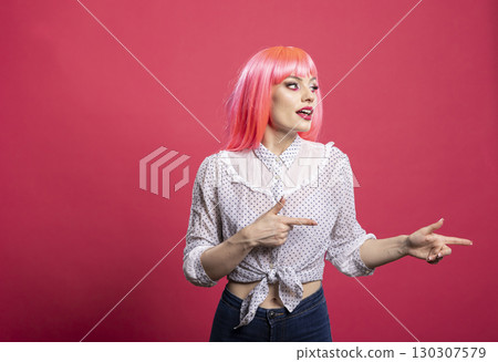 Excited woman with pink wig stands isolated on pink background, smiling and pointing finger towards blank copy space. Energetic and vibrant model presenting area for advertisement or promotional Excited woman with pink wig stands isolated on pink background, smiling and pointing finger towards blank copy space. Energetic and vibrant model presenting area for advertisement or promotional 130307579