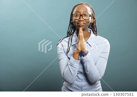 African American woman with palms together, looking at camera and pleading. Desperate black female individual standing with hands clasped, praying, hoping for good news in blue background studio. African American woman with palms together, looking at camera and pleading. Desperate black female individual standing with hands clasped, praying, hoping for good news in blue background studio. 130307581