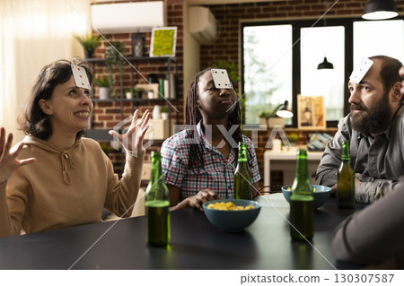 Group of young, diverse friends enjoying games and snacks in apartment. Caucasian and black people with cards on forehead, playing guessing game at table with beer and chips during weekend hangout. 130307587