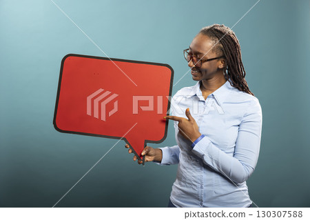 Cheerful woman holding and looking at red cardboard sign, standing against isolated background. Smiling black female individual in blue shirt grasping blank speech bubble, ideal of advertisement. 130307588