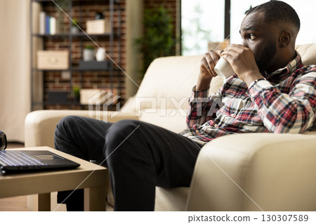 Young black entrepreneur sits on couch, sipping coffee and taking mindful break from remote work duties. African american man gazes thoughtfully around cozy living room during peaceful afternoon pause Young black entrepreneur sits on couch, sipping coffee and taking mindful break from remote work duties. African american man gazes thoughtfully around cozy living room during peaceful afternoon pause 130307589