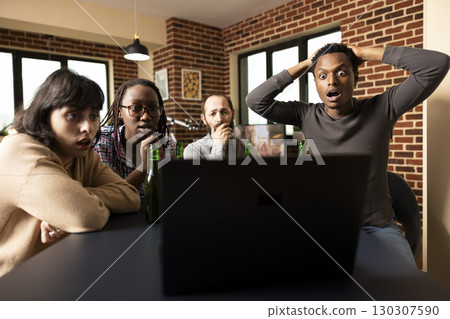 Shocked black man with hands on head, watching online content with friend, gathered around table in brick wall room. Young diverse adults reacting with wide eyes to suspenseful scene on laptop screen. Shocked black man with hands on head, watching online content with friend, gathered around table in brick wall room. Young diverse adults reacting with wide eyes to suspenseful scene on laptop screen. 130307590