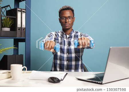 Male freelance sitting at the table and holding dumbbells towards the front, doing small exercises during work. African american man with glasses, using hand weights and looking at camera. Male freelance sitting at the table and holding dumbbells towards the front, doing small exercises during work. African american man with glasses, using hand weights and looking at camera. 130307598