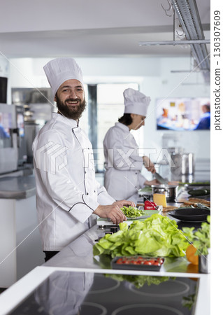 Professional chef in a modern kitchen preparing gourmet dishes with fresh produce, using culinary tools and stainless steel utensils. Male cook working in the gastronomy industry. 130307609