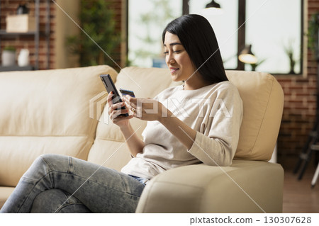 Cheerful female entrepreneur sitting on couch in modern apartment, using smartphone and debit card for digital payment. Asian businesswoman completing online purchase on mobile device. 130307628