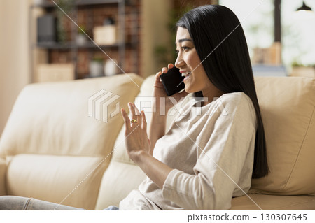 Smiling businesswoman talks on mobile device with investor, enjoying productive conversation. Cheerful female freelancer sitting on sofa with phone to ear, having discussion with potential client. 130307645