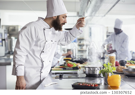 Professional chef in uniform tasting a savory sauce from a pan on the stove, ensuring the perfect balance of flavors. Delicious dish simmers on the stovetop at a modern restaurant kitchen counter. Professional chef in uniform tasting a savory sauce from a pan on the stove, ensuring the perfect balance of flavors. Delicious dish simmers on the stovetop at a modern restaurant kitchen counter. 130307652