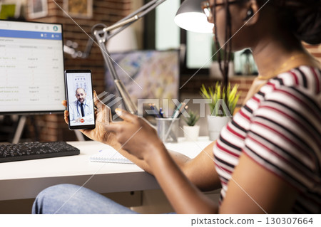 Black woman holding cellphone and having discussion with professional doctor on video call. African american female individual sharing health updates with caucasian physician during virtual meeting. Black woman holding cellphone and having discussion with professional doctor on video call. African american female individual sharing health updates with caucasian physician during virtual meeting. 130307664