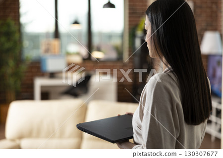 Self employed asian woman walking into brick wall living room with laptop in hand. Female entrepreneur standing and holding digital device, planning to work from home, researching marketing strategies 130307677