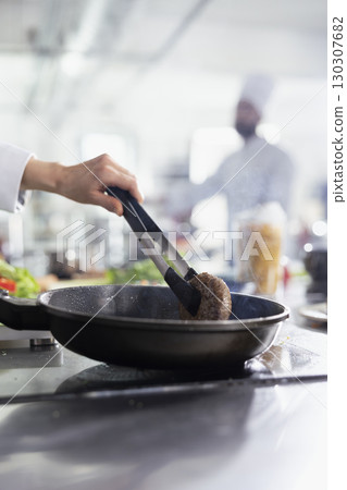 Female chef preparing a juicy beef steak piece for a gourmet meal on the stove, flipping the raw meat with tongs in the pan. Culinary expert preparing a fillet in oil, gastronomy. Close up. 130307682