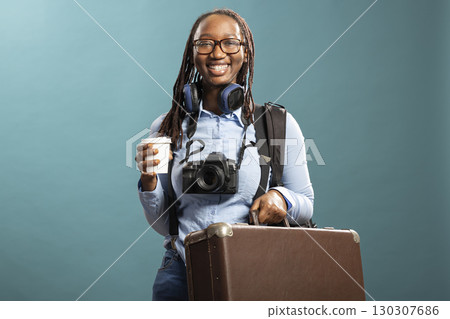 Joyful female traveler poses with coffee and DSLR, wireless headphones around neck, standing confidently in front of blue background. Happy photographer carrying suitcase and camera, ready for trip. 130307686