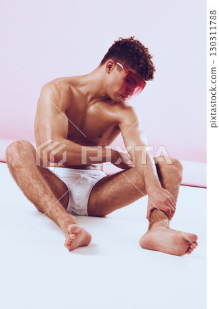 Stylish young man with curly hair sitting on the floor, wearing pink sunglasses and white underwear, posing against a soft pink background. Fashion and self-expression concept. 130311788