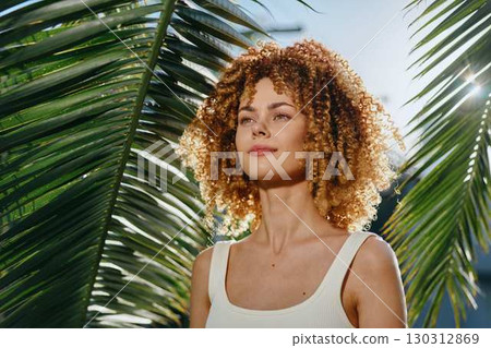 young woman with curly hair stands confidently under bright sunlight surrounded by tropical palm leaves, wearing a white tank top with natural makeup. young woman with curly hair stands confidently under bright sunlight surrounded by tropical palm leaves, wearing a white tank top with natural makeup. 130312869