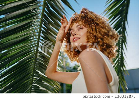 Smiling woman with curly hair enjoys sunlight under palm leaves, wearing white sleeveless top, vibrant outdoor summer scene with clear blue sky background. 130312873