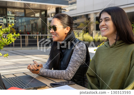 Two businesswomen working remotely on a laptop and smiling outdoors Two businesswomen working remotely on a laptop and smiling outdoors 130313593