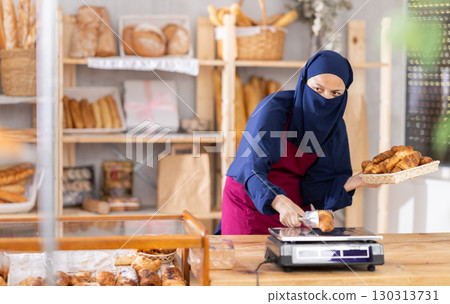Asian Muslim woman seller in hijab weighs croissants on scale, 130313731