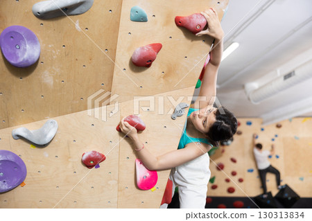 Asian female client of fitness room trains doing exercises on artificial wall for climbing, top view Asian female client of fitness room trains doing exercises on artificial wall for climbing, top view 130313834
